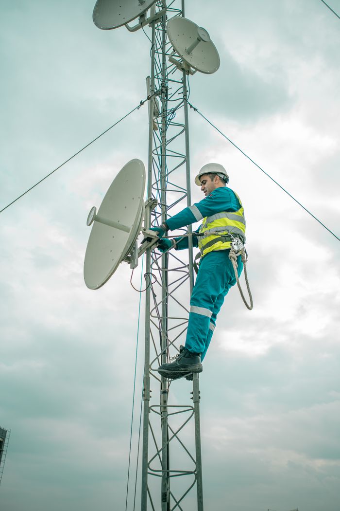A worker in safety gear installs a satellite dish on a telecom tower under an overcast sky.