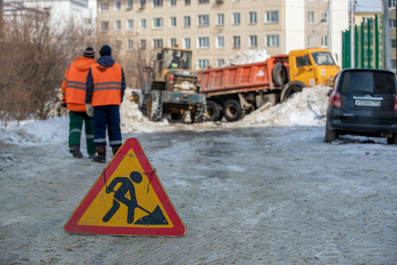 Winter street construction with workers and vehicles in a snowy city setting.